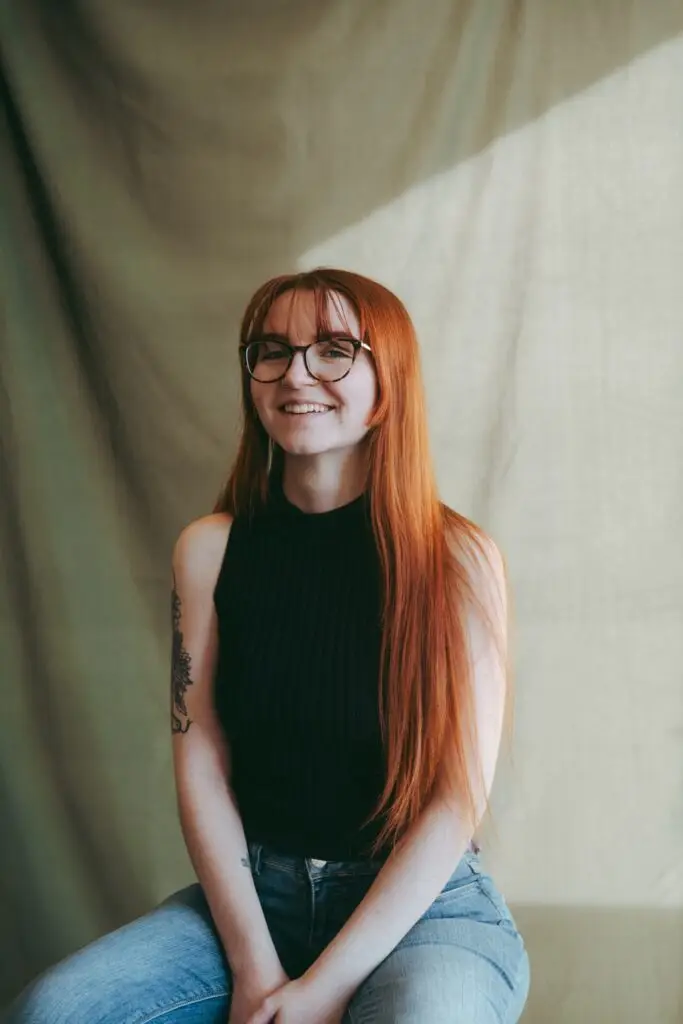 A red-headed woman sitting on a stool against a green backdrop, smiling.