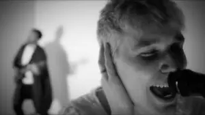 A black and white image of a man singing close to the camera, another man playing guitar in the background.