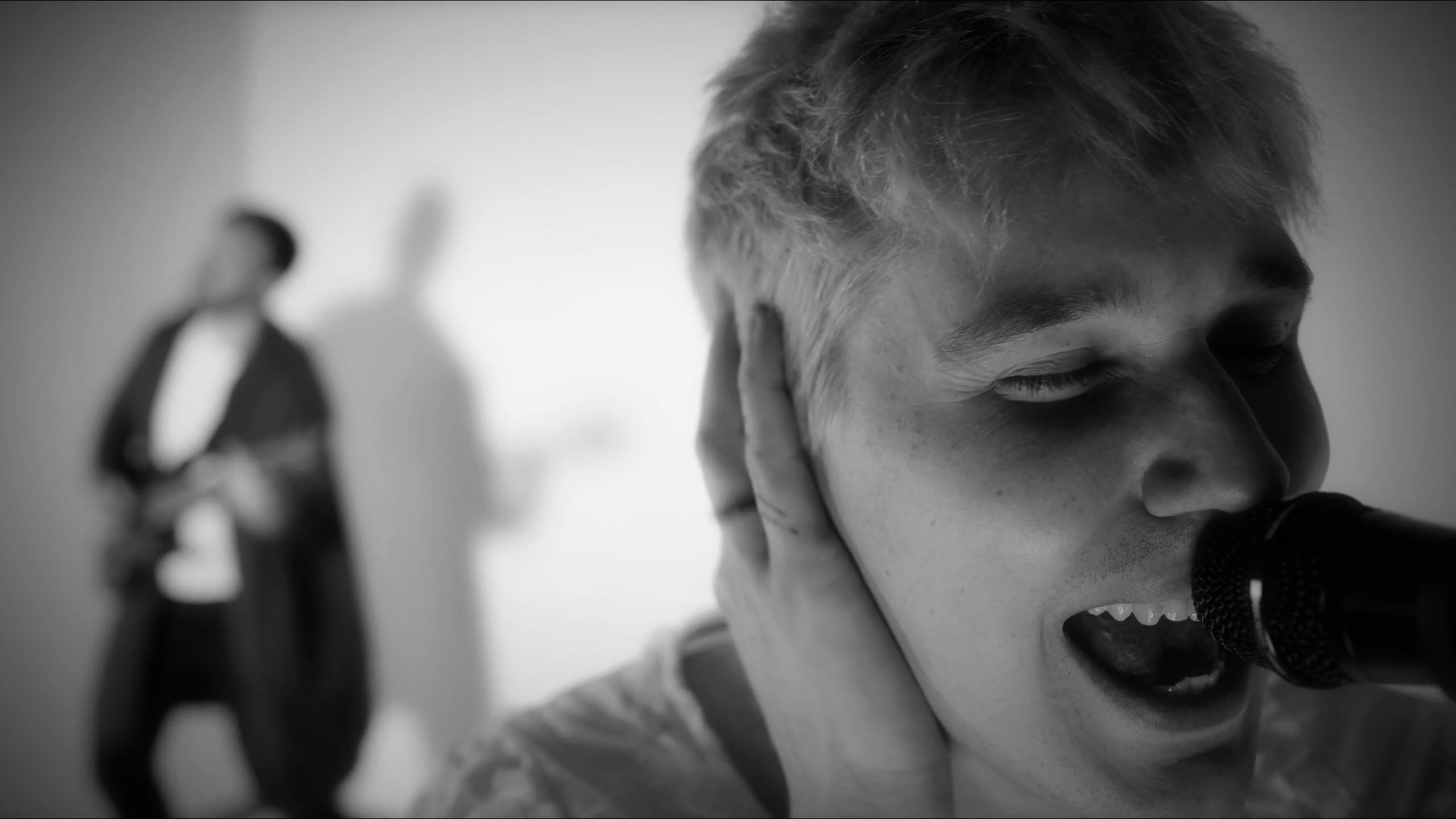 A black and white image of a man singing close to the camera, another man playing guitar in the background.